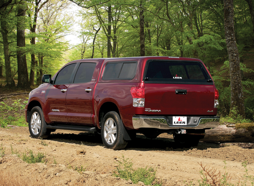 A maroon pickup truck with a canopy is parked on a dirt trail in a forested area. Lush green trees surround the vehicle, and the ground is uneven with patches of dirt and grass. The trucks back is facing the viewer.