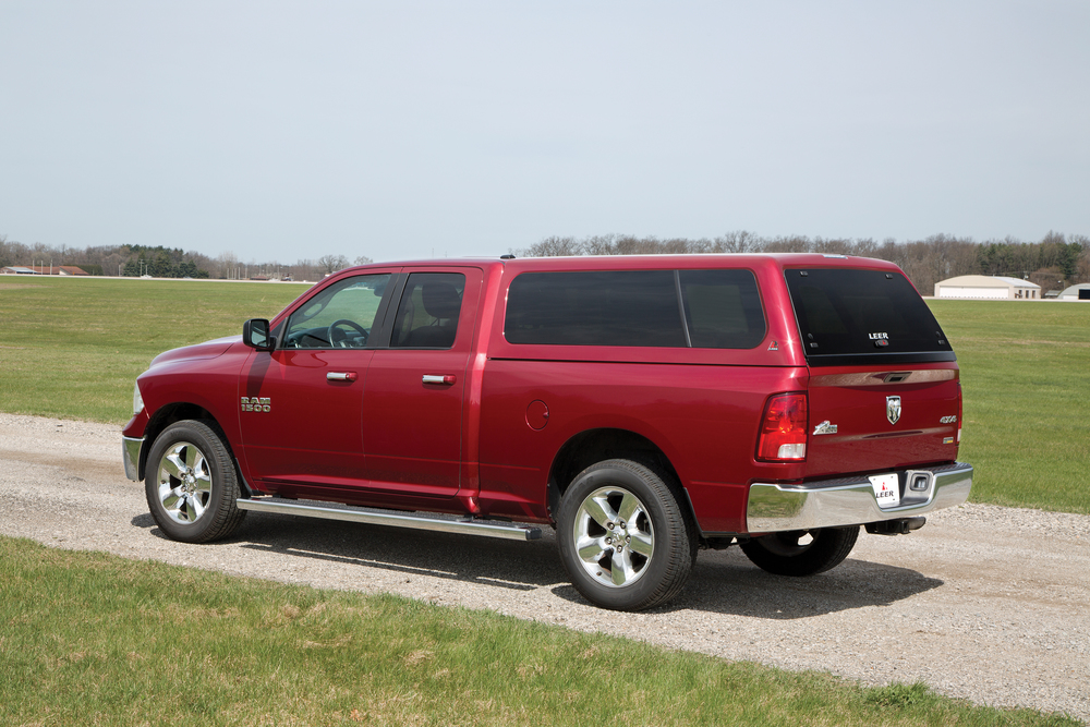 A red pickup truck with a canopy is parked on a gravel path beside a grassy field. Trees and a few buildings are visible in the background under a clear sky.