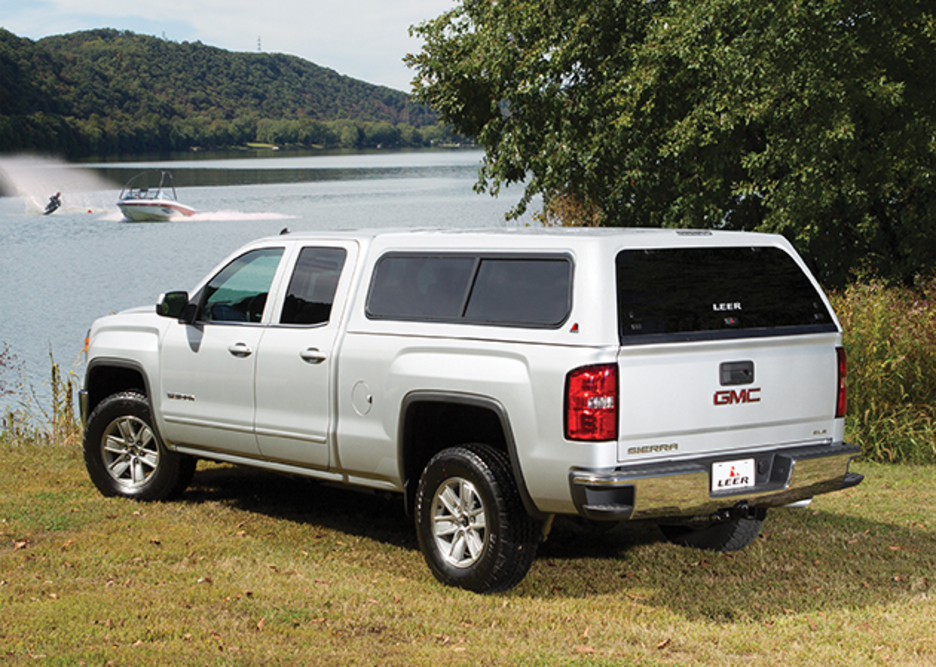 A silver GMC Sierra pickup truck with a camper shell is parked on grass by a lake. In the background, a boat is speeding across the water, creating a wake. Trees and lush foliage surround the lake, under a clear sky.