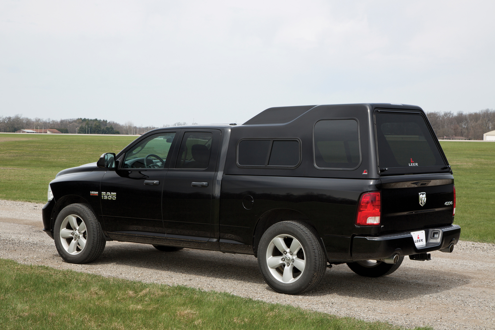 A black pickup truck with a covered cargo bed is parked on a gravel road beside a grassy field. The truck has a cap mounted on the back, and there are trees in the distant background under a cloudy sky.