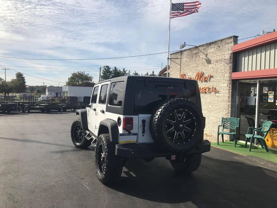 A white Jeep Wrangler with large black wheels and tires is parked in a lot in front of a Pen Mar Sales building. 