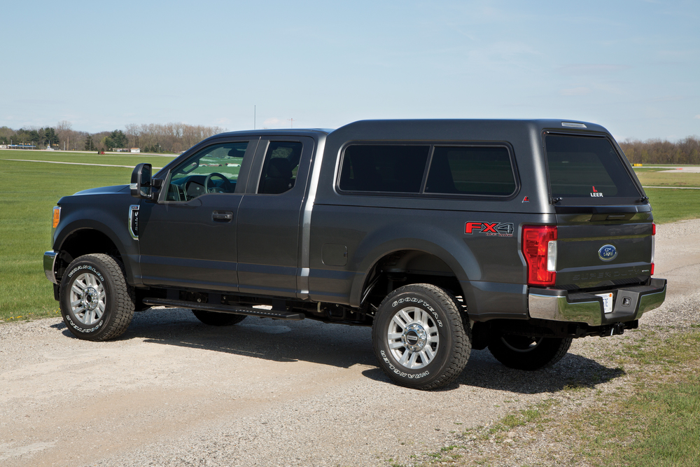 A dark gray pickup truck with an extended cab and a windowed camper shell on the back is parked on a gravel path, surrounded by grassy fields under a clear blue sky.