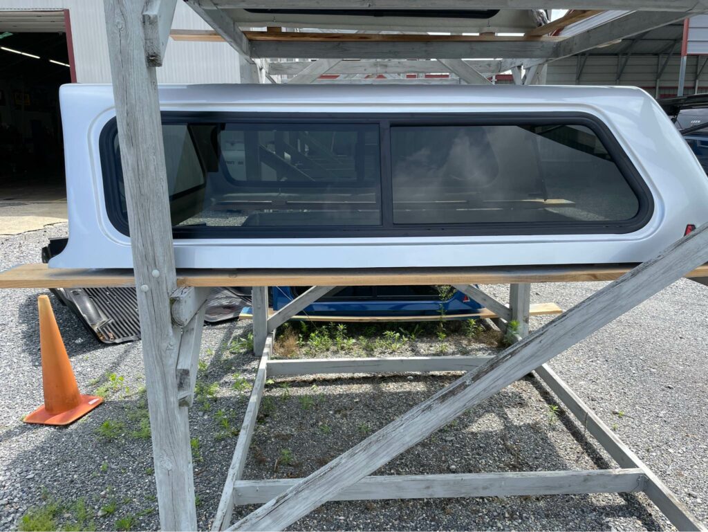 A white truck bed topper with tinted windows is displayed on a wooden rack. It is positioned outdoors on a gravel surface near an industrial building.