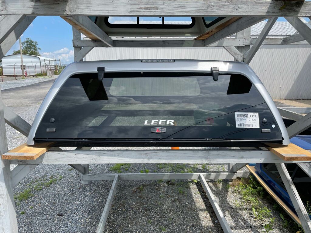 A silver and black truck bed cap with a LEER logo is mounted on a wooden rack outdoors. The cap has a large rear window and is supported by a metal frame. A vehicle and building are visible in the background.