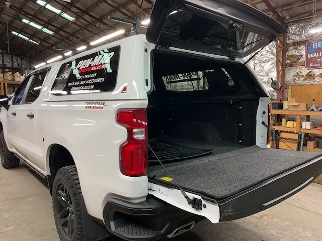 A white pickup truck with an open camper shell and tailgate is shown in a garage. The interior of the truck bed is lined with carpet. The garage has various tools and equipment, with a sign partially visible on the wall.