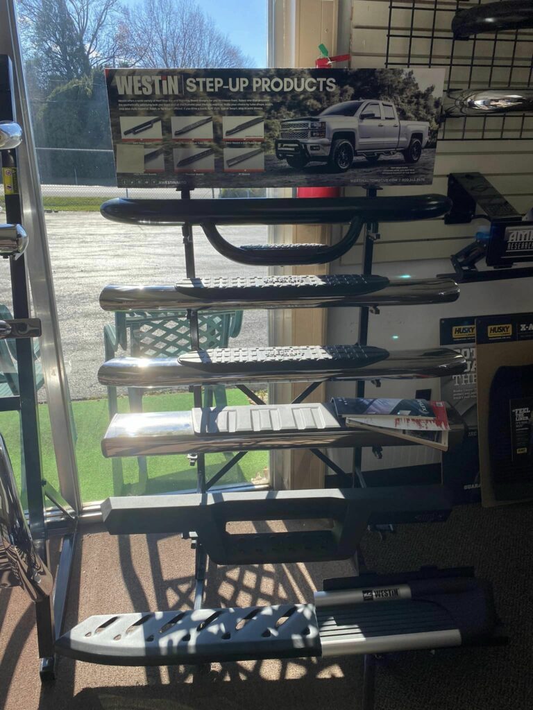 Display of several truck step bars in a store. A banner above shows a truck with Westin Step-Up Products branding. Sunlight streams through windows, casting shadows on the floor. Various brochures and product information are on the shelves.