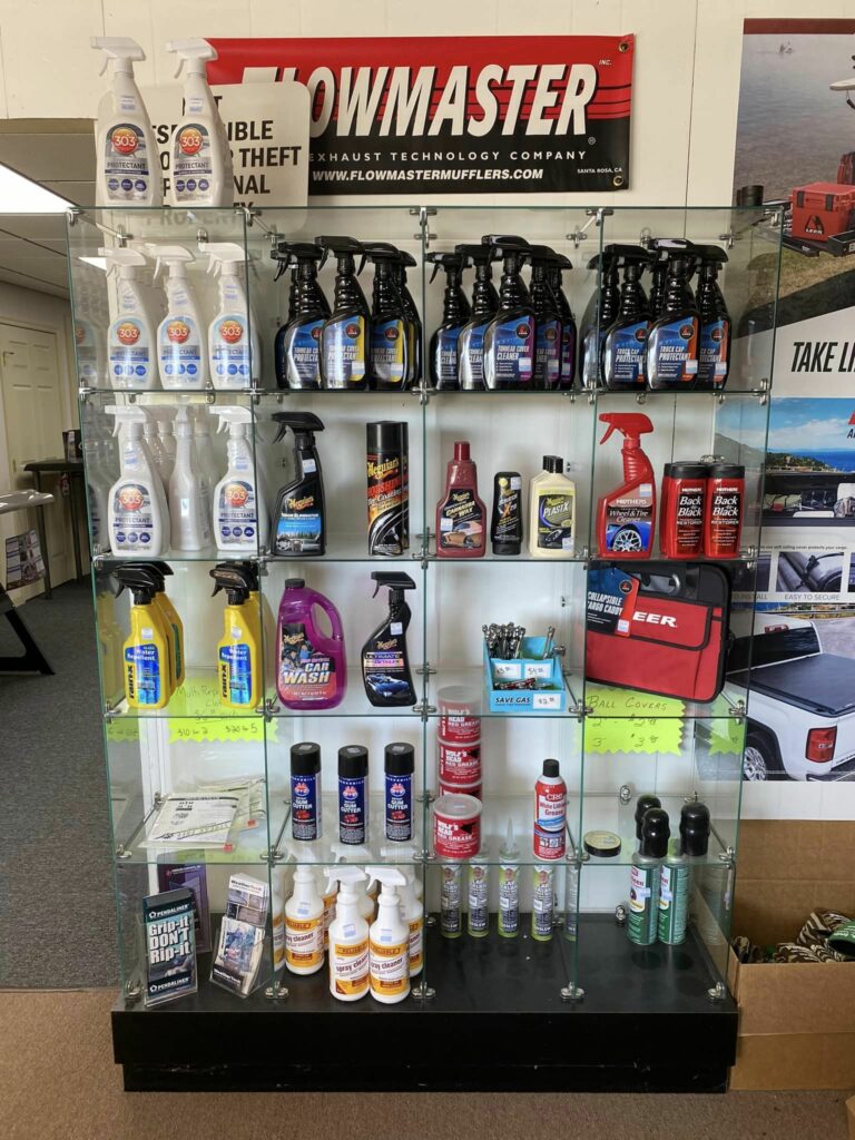 A display case filled with various automotive cleaning and maintenance products, including spray bottles and cans. Above the case is a Flowmaster sign. The surrounding walls have automotive-themed posters and advertisements.