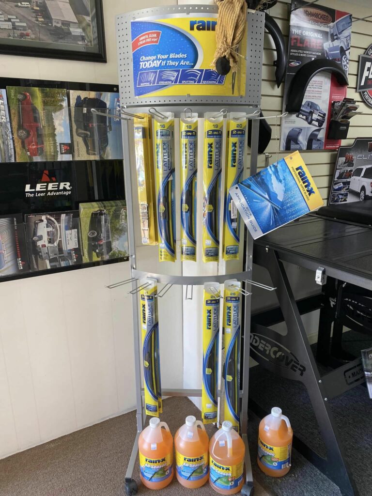 A display stand in a store holds multiple Rain-X wiper blades and bottles of Rain-X cleaning fluid. A poster with information about the product is attached to the top, and various automotive posters are visible on the walls in the background.