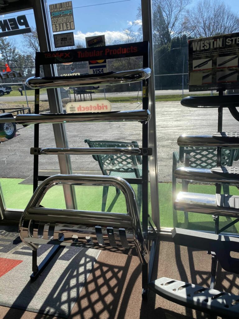 Display rack with chrome vehicle accessories inside a store. Sunlight streams through the large window, casting shadows on the floor. Green outdoor chairs and a fence are visible outside. Various product logos are on the rack.