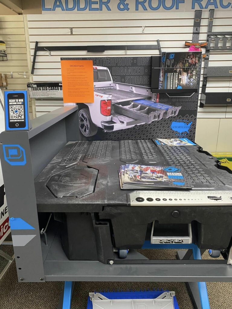 Display of a truck bed storage system with promotional materials. The setup includes sample drawers and a QR code sign. A large image of a white pickup truck with an installed storage system is in the background.