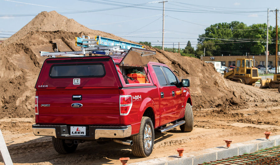 A red Ford pickup truck with a canopy and ladder on top is parked on a construction site. There is a pile of dirt in the background, and various construction materials and equipment are visible around the area.