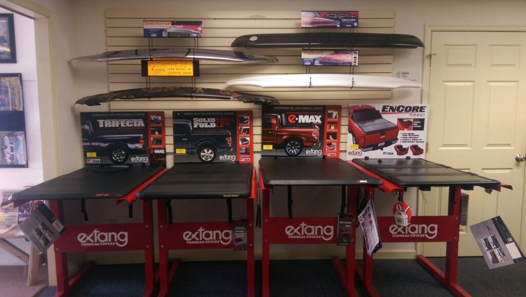 A store display featuring various truck bed covers by Extang. The products include models like Trifecta, Solid Fold 2.0, and e-Max, with miniature truck models on top. The backdrop has mounted sample covers.