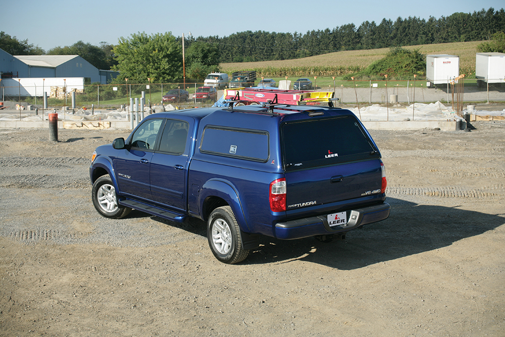 A blue pickup truck with a canopy is parked on a dirt construction site. The truck has a ladder on top and is near foundations and trees. There are buildings in the background.