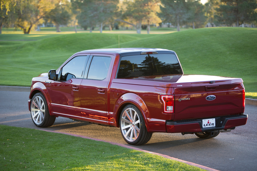 A shiny red pickup truck with tinted windows is parked on a paved road next to a grassy area. It's a sunny day, and trees are visible in the background.